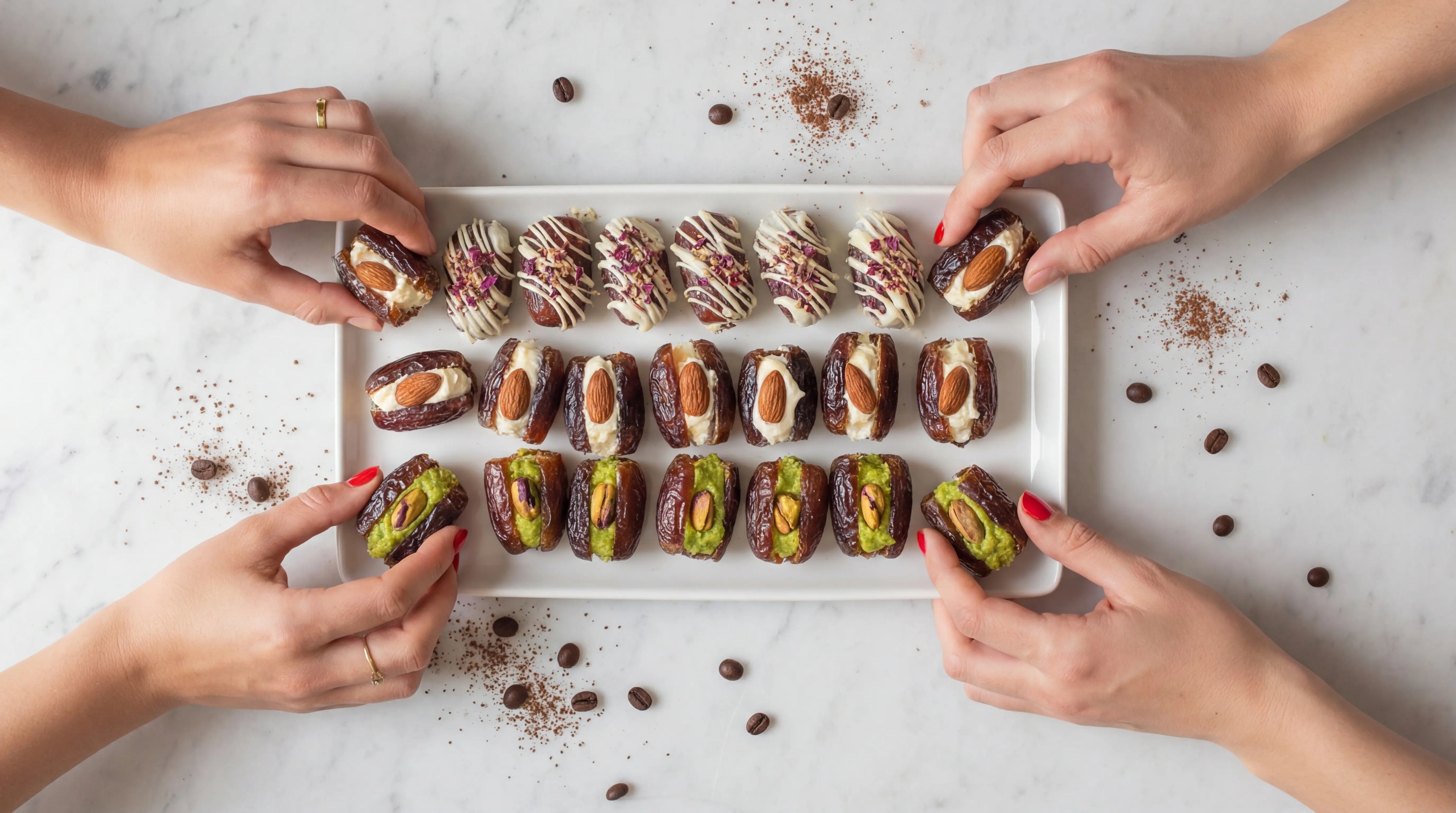 Four hands reaching towards a tray of assorted dried fruits and nuts on a light surface.